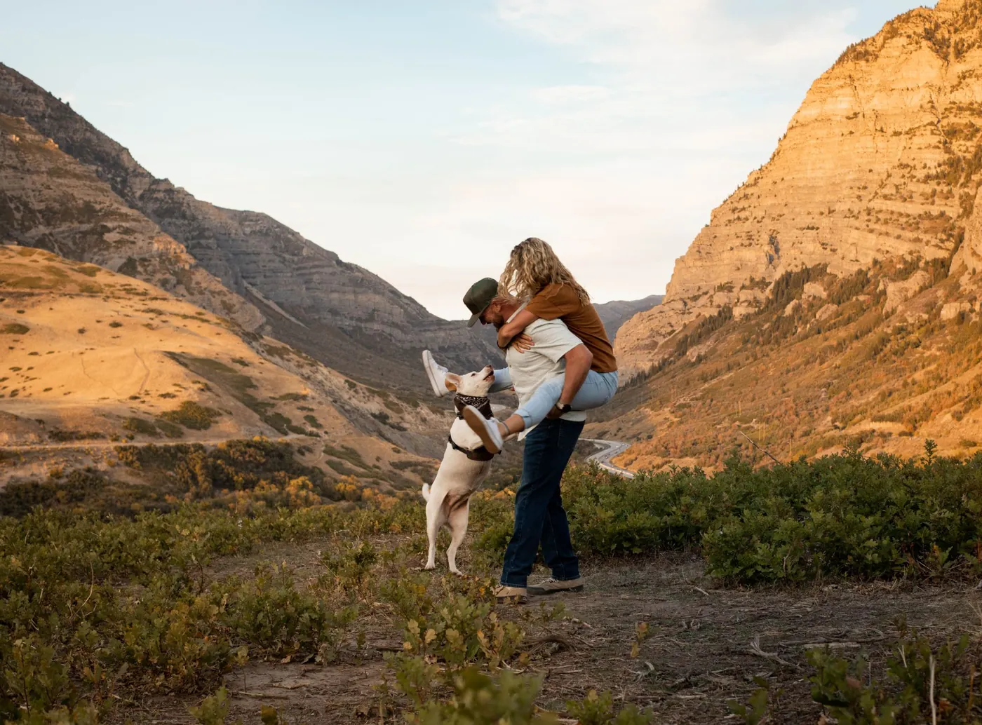 James Lewis carrying Kate as their dog, Mattie, jumps up to join in, in a mountain canyon